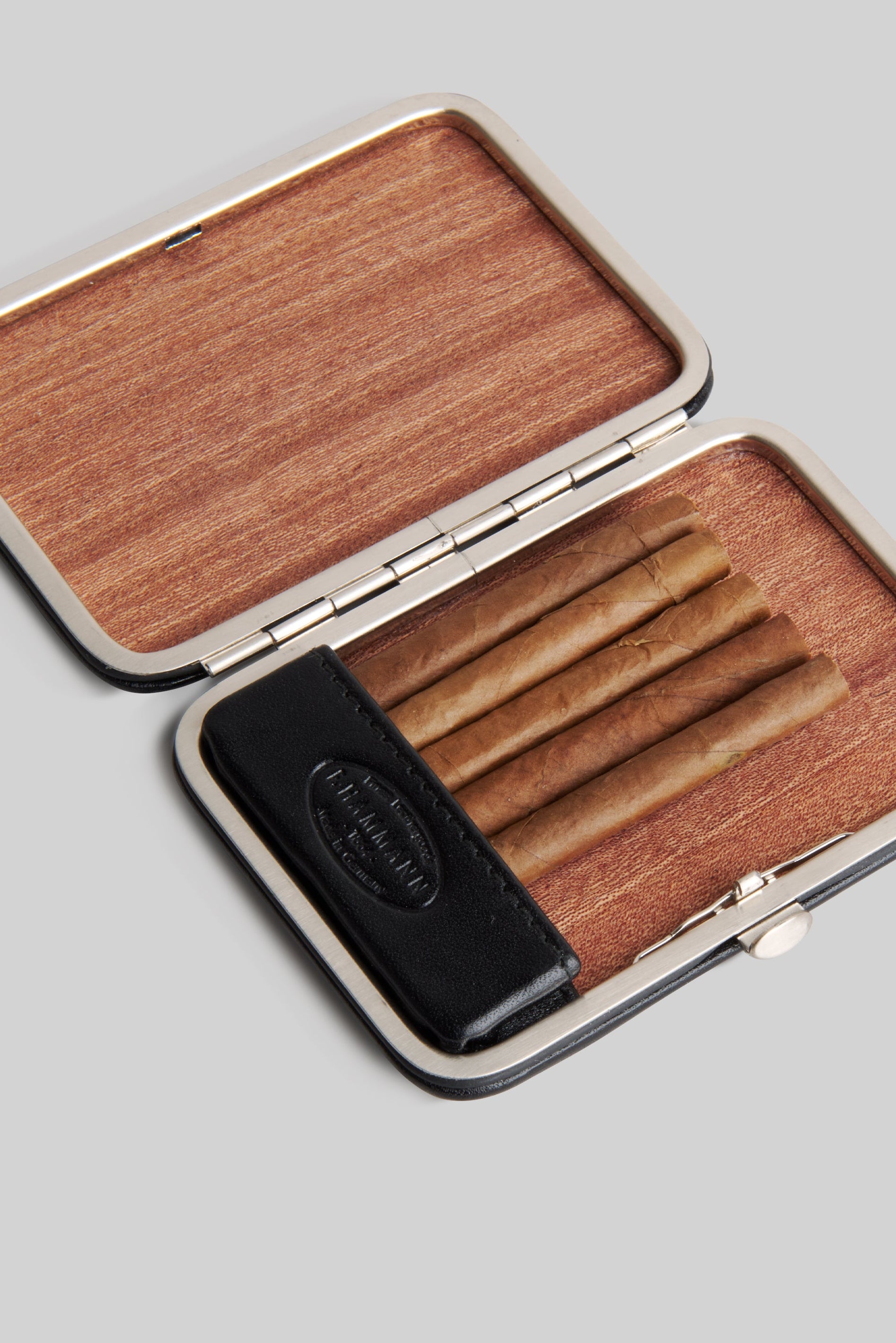 An open rectangular cigar case with a wood-grain patterned interior, silver-toned metal edges, and a black leather insert holding several cigars, shown on a light gray background.